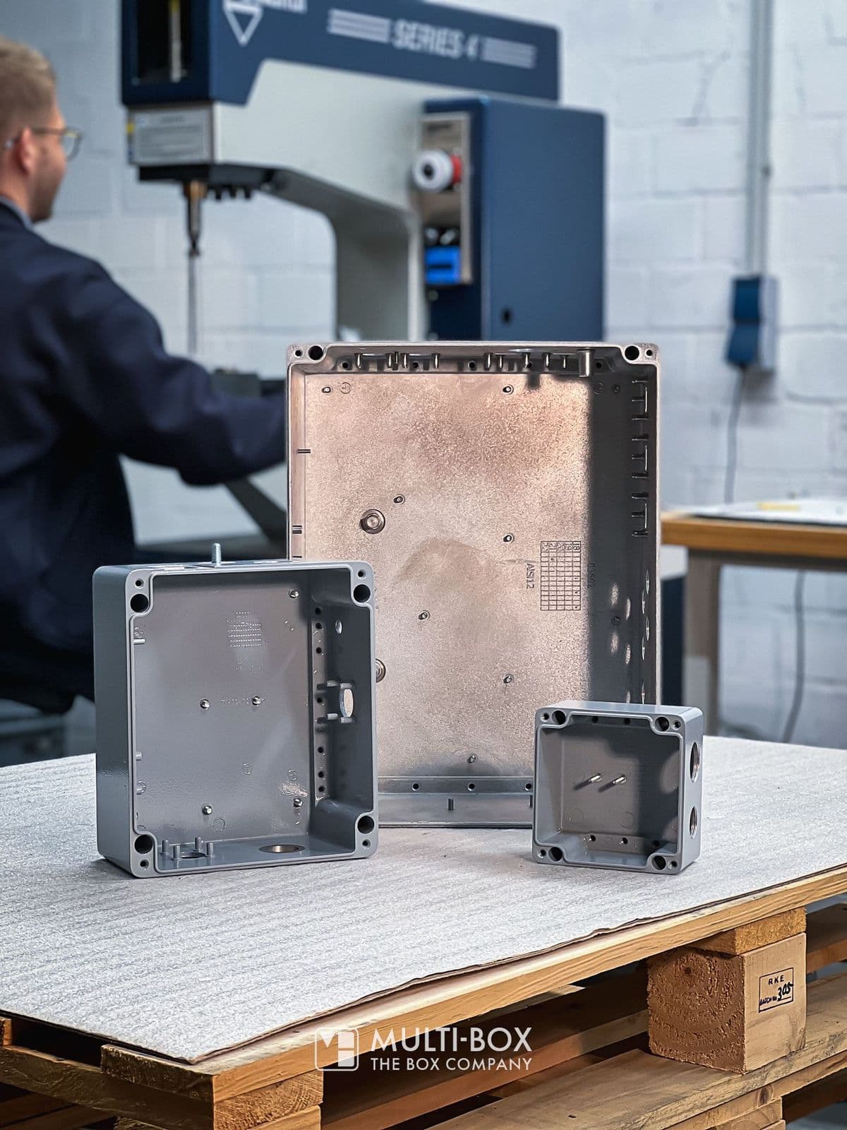 Drei Aluminiumgehäuse in verschiedenen Größen auf einer Werkbank neben einer Bolzen-Einpressmaschine.