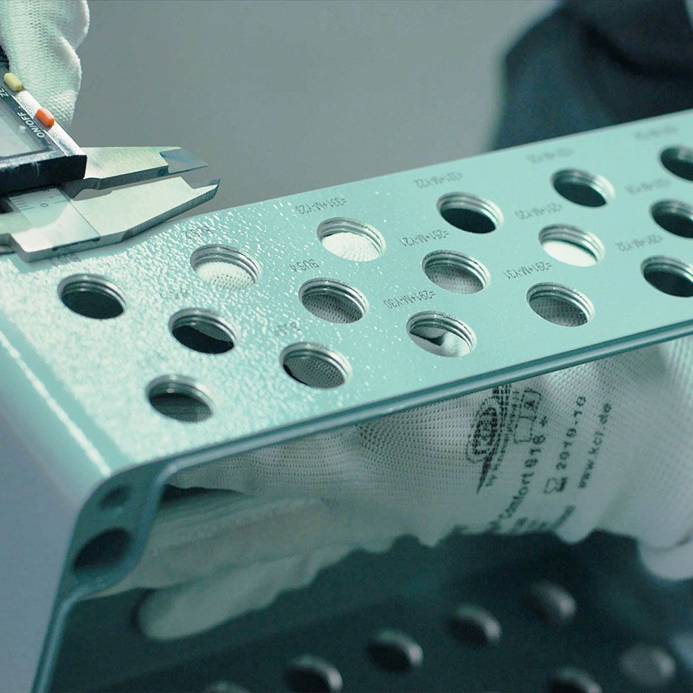 A perforated metal part is held by a hand wearing a work glove.
